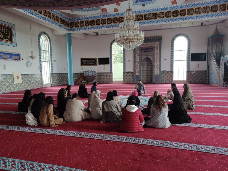 leerlingen zittend op de grond tijdens een bezoek aan een moskee waar ze uitleg krijgen van de imam