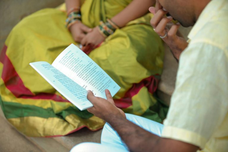 Photo of an Indian Man Reading a Book while Sitting on a Ground with a Woman in Traditional Clothing in the Background