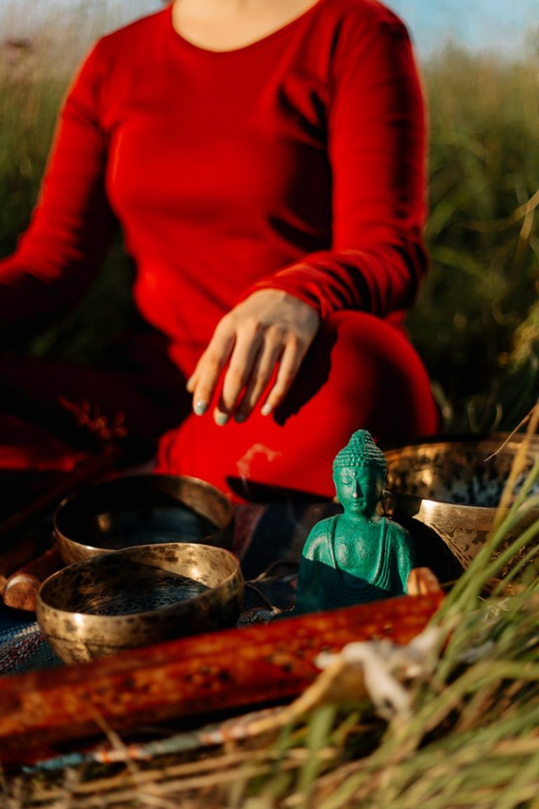 Woman in red meditating outdoors with Tibetan singing bowls and a Buddha statue.